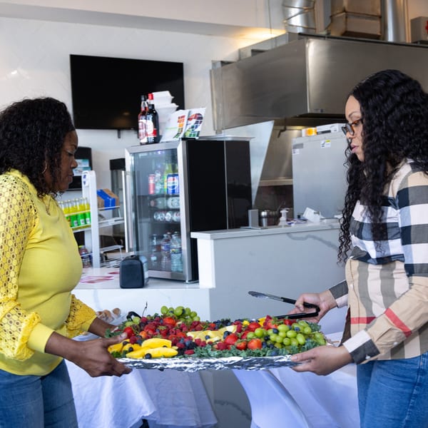 Two women holding a large platter of fresh fruit, including grapes, berries, and pineapple.