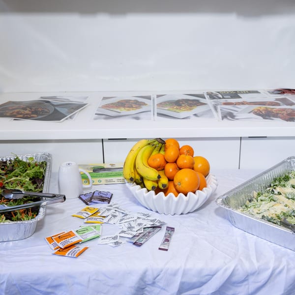 A buffet table with salads, a fruit bowl, tea bags, and coffee packets, with food photos in the background.