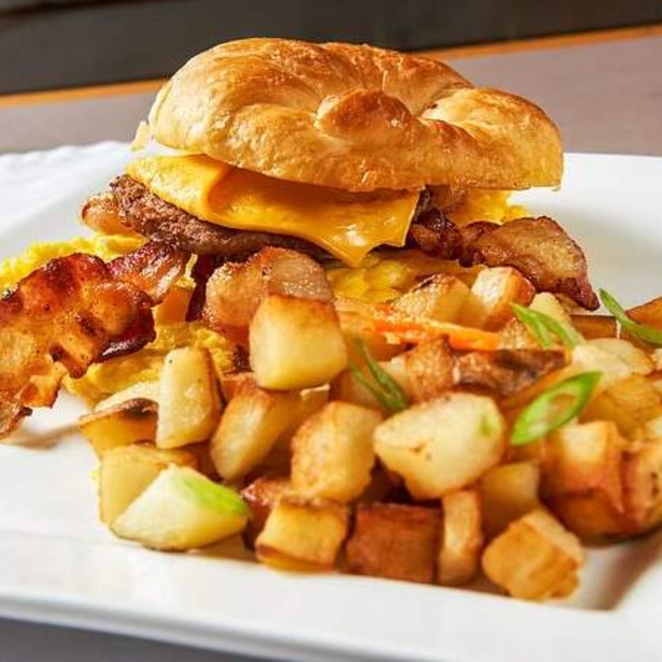 A breakfast sandwich on a croissant with egg, cheese, sausage, bacon, and a side of home fries.