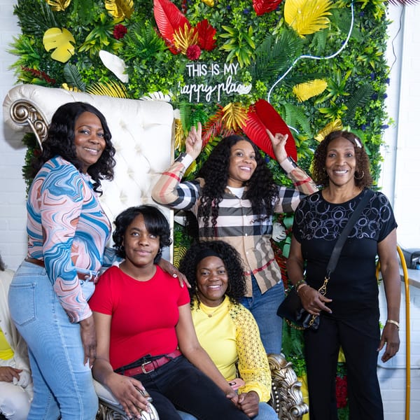 Five women smiling in front of a green wall with tropical leaves and a "This is my happy place" sign.