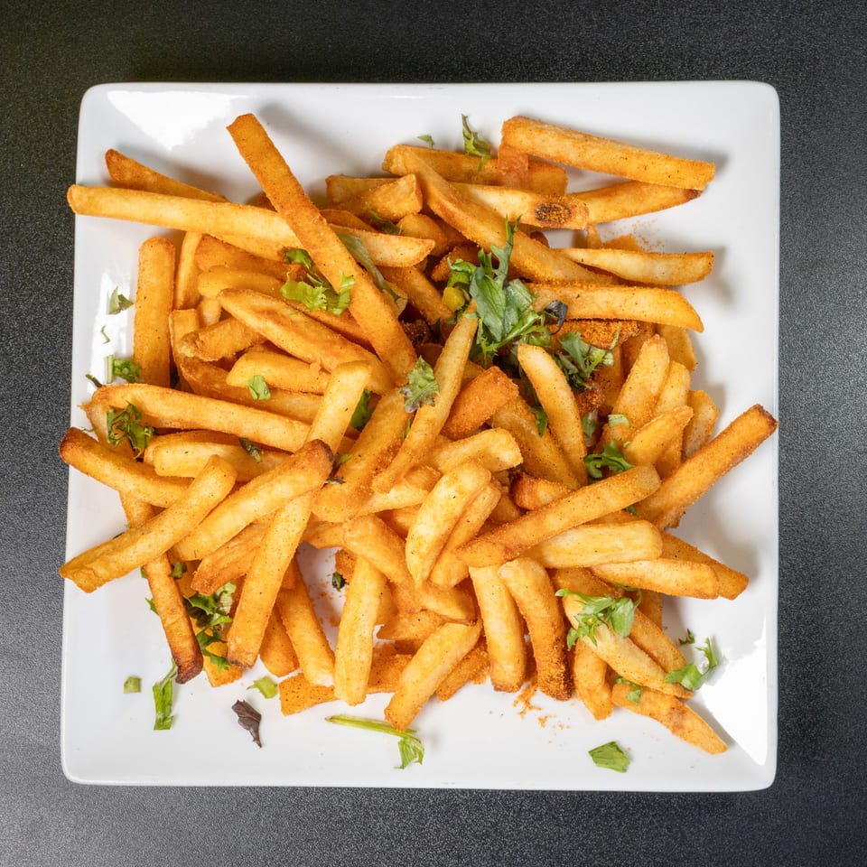 A white rectangular plate filled with golden-brown, seasoned French fries and green herbs.