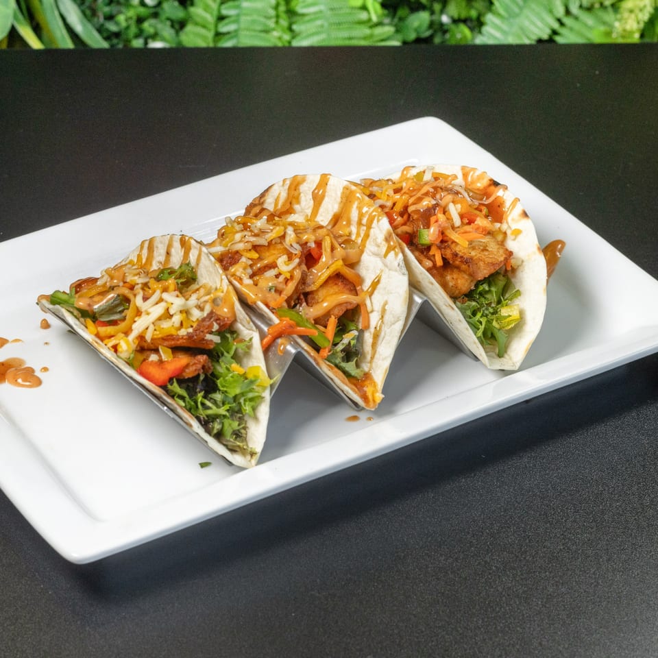 Three tacos in metal holders, filled with meat, lettuce, shredded cheese, and sauce on a white plate.