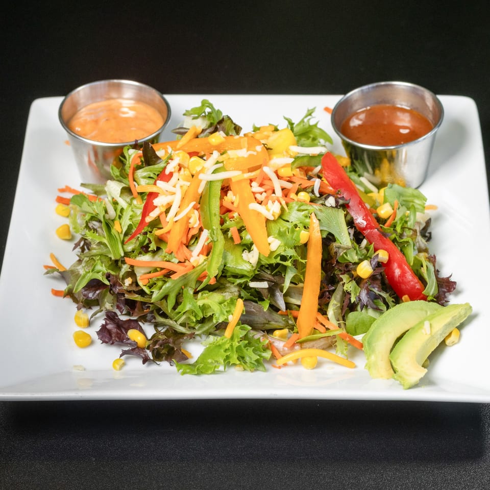 Colorful garden salad with mixed greens, bell peppers, corn, shredded cheese, avocado, and two dressings.