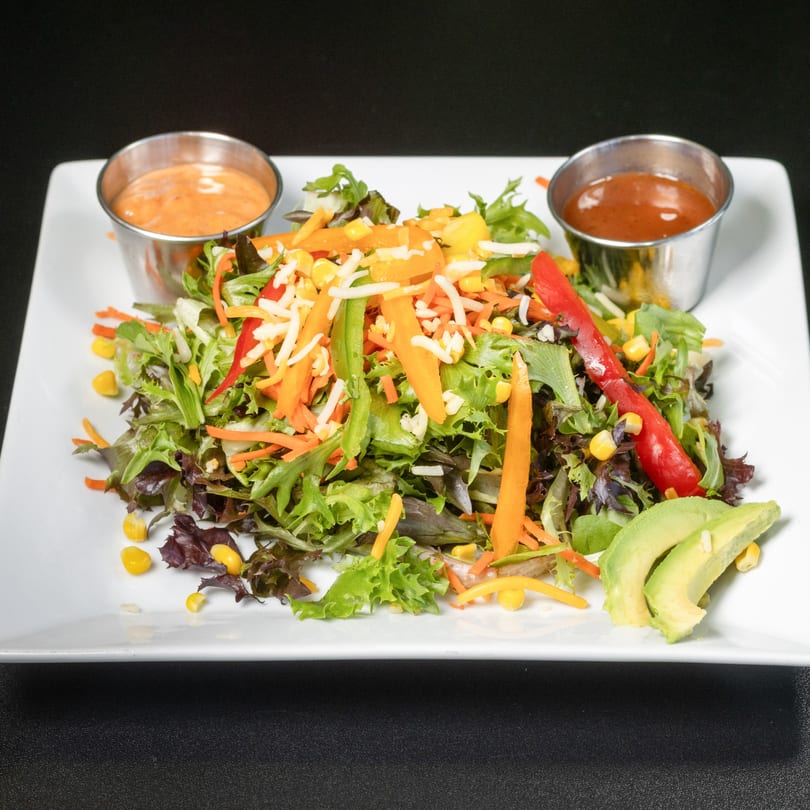 Colorful garden salad with mixed greens, bell peppers, corn, shredded cheese, avocado, and two dressings.